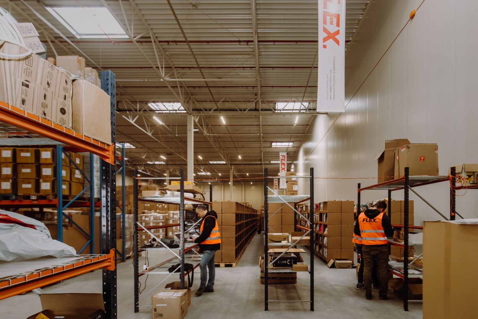 Workers in orange vests organize shelves in a spacious warehouse filled with boxes. The scene reflects orderliness and efficiency.