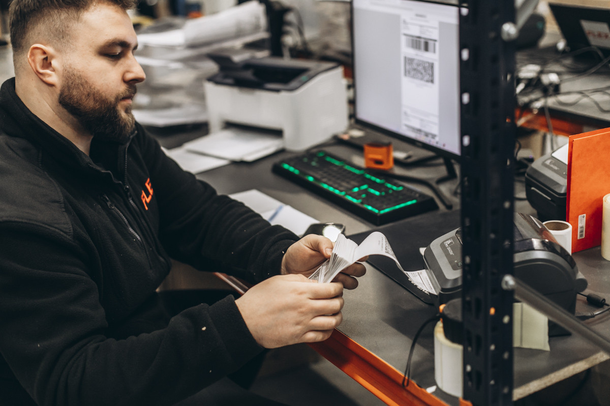 FLEX. warehouse employee reviewing printed shipping labels at a dispatch workstation equipped with label printers and a computer monitor.