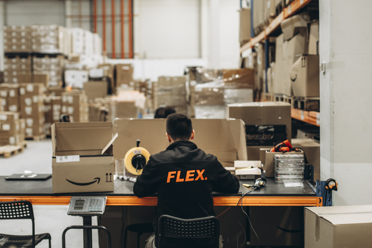 FLEX. warehouse employee working at a packing station with shipping supplies, cartons, and warehouse inventory visible throughout the fulfillment facility.