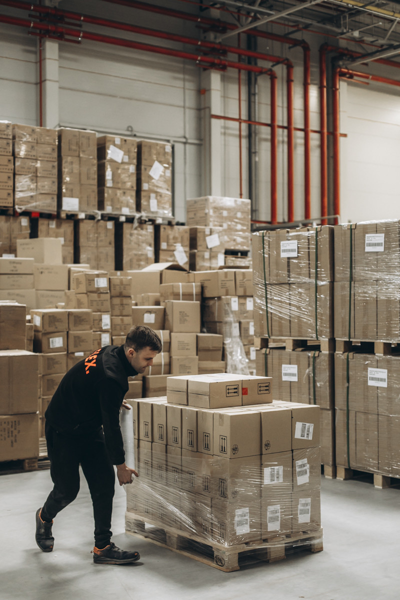 FLEX. warehouse employee securing a pallet of cartons with stretch wrap in a warehouse distribution and storage area surrounded by palletized inventory.