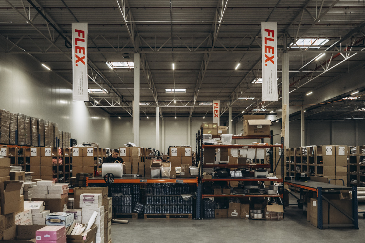 Wide view of a fulfillment warehouse with packing stations, storage shelving, palletized inventory, and FLEX. branding banners suspended above the work area.
