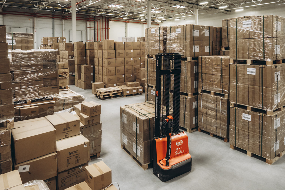 Wide view of a warehouse storage area with palletized cartons, shrink-wrapped inventory, and electric material handling equipment positioned between storage locations.