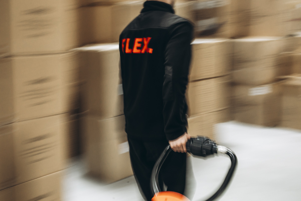 FLEX. warehouse worker moving through a fulfillment facility with pallet handling equipment, surrounded by stacked cartons and inventory storage.